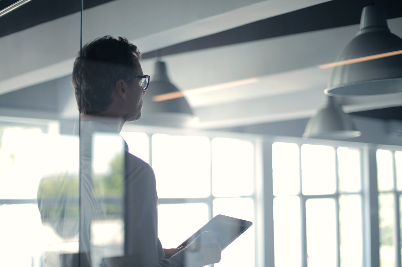 our-story A businessman holding a tablet in an office, looking thoughtfully through a glass wall.